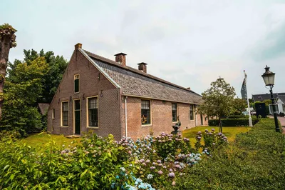 Een oud, karakteristiek huis met een rieten dak, omgeven door groene planten en bloemen.