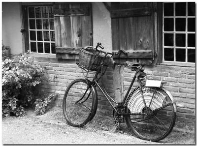 Zwarte en witte foto van een oude fiets met een mand, leunend tegen een muur met houten ramen.