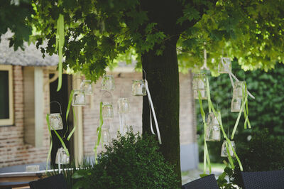 Decoratieve glazen lantaarns hangen aan een boom, omringd door groene bladeren en een tuinsetting.