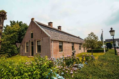 Een oud, karakteristiek huis met een rieten dak, omgeven door groene planten en bloemen.